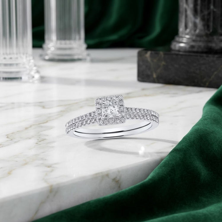 The 14k Gold Princess Diamond Square Halo Wedding Ring Set shines with a striking princess diamond at its center, surrounded by a square halo and paired with a dazzling diamond band. Displayed gracefully on a marble surface, the ring set is beautifully framed by white columns and lush green velvet fabric in the background.