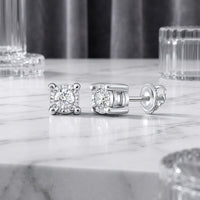 A pair of 10k Gold Round Diamond Stud Earrings 1/6 Cttw with screw-back settings rest on a white marble surface, their brilliance enhanced by expert craftsmanship, with clear glass decor and a gray curtain in the background.