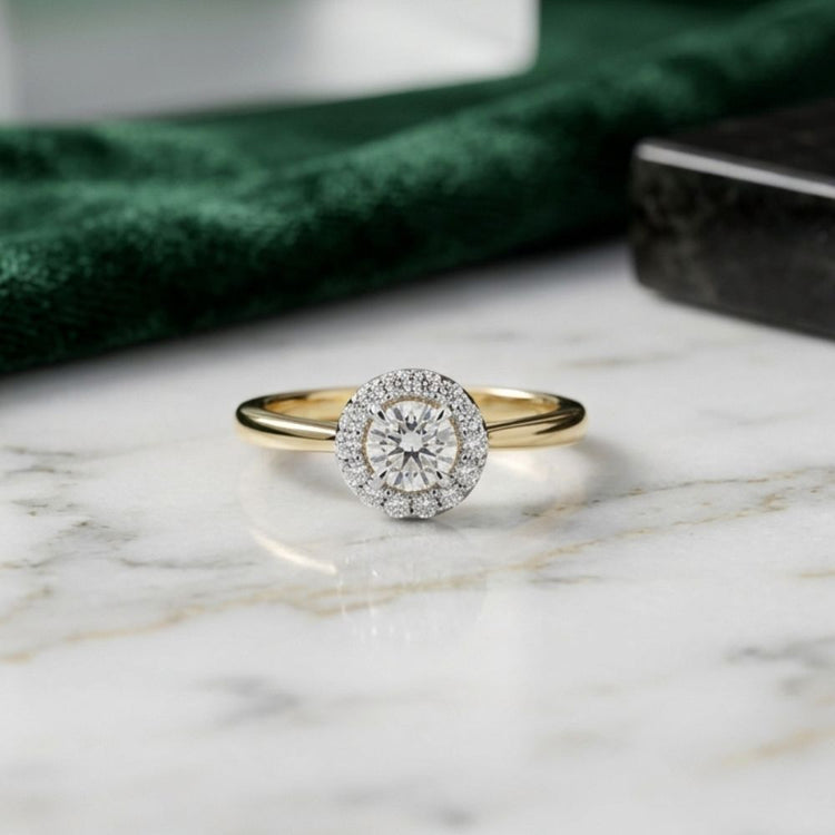 The 14k Gold Round Diamond Halo Engagement Ring, showcased on a marble surface, features a brilliant round diamond center stone encircled by a sparkling halo of smaller diamonds—an elegant and timeless choice for bridal engagement.