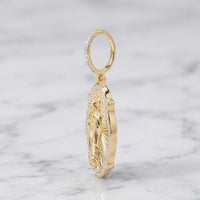 The Diamond Virgin Prayer Charm 3/8CT in 10K gold features an engraved religious figure with sparkling white stones on the border and loop, displayed upright on white marble—a bold pendant for men.