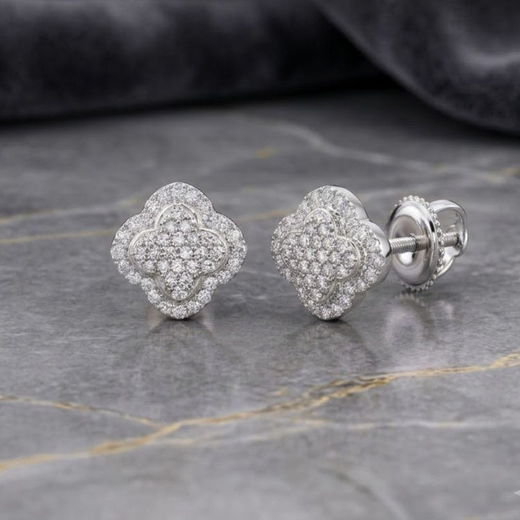 A pair of 1/5 CTTW Diamond Clover Earrings in 10K Yellow & White Gold, featuring a clover-shaped stud adorned with sparkling diamonds and secure screw-back posts, displayed on a gray marble surface against a dark fabric background.