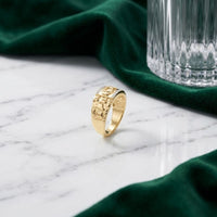 A 10k Gold Dad Men’s Ring with Natural Diamonds featuring an engraved design stands upright on a white marble surface, surrounded by green velvet fabric and a clear glass with a textured pattern.