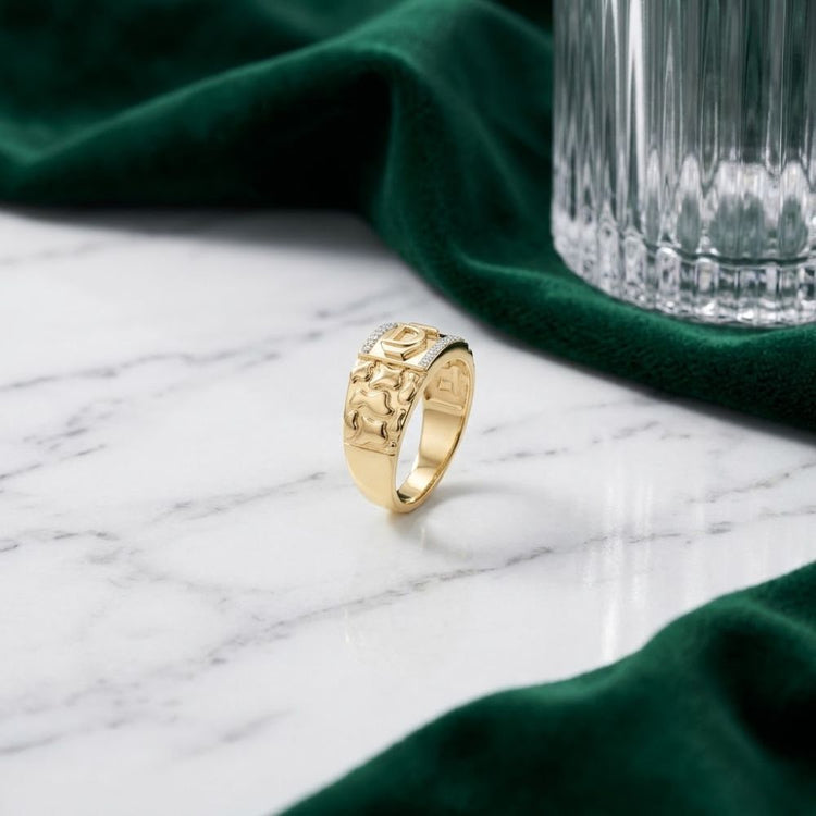 A 10k Gold Dad Men’s Ring with Natural Diamonds featuring an engraved design stands upright on a white marble surface, surrounded by green velvet fabric and a clear glass with a textured pattern.