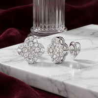 The 1/6 CTW Natural Diamond Clover Earrings in 10K Gold are displayed on a white marble surface, set against a backdrop of a glass column and rich red velvet fabric.