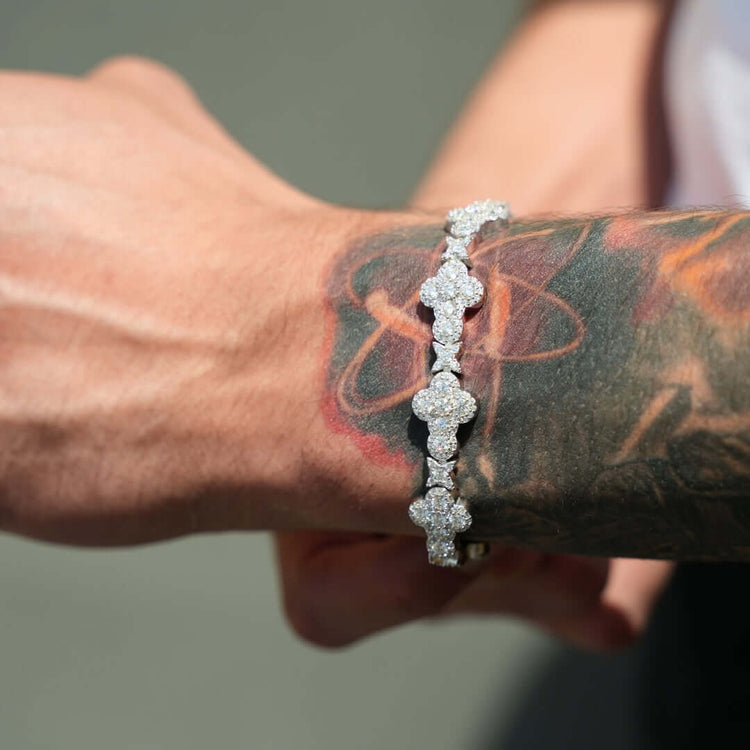 A close-up of a wrist shows a sparkling Moissanite Cross Fancy Bracelet in 925 sterling silver. The forearm features a vibrant butterfly tattoo in red, orange, and black ink. The background is softly blurred.
