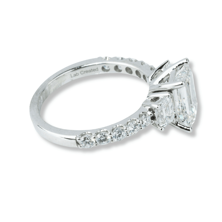 The 3.8 CTW 14k White Gold Three Stone Emerald Engagement Ring features a large emerald-cut diamond with two smaller lab-grown diamonds, round accents, and "Lab Created" engraved inside the band.