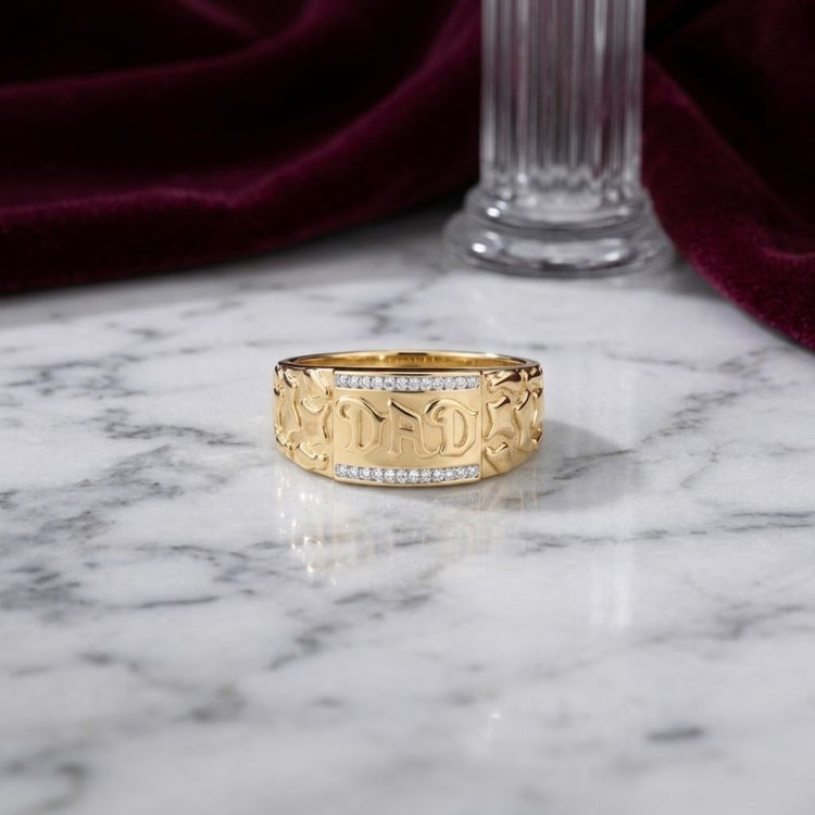 A 10k Gold Dad Men’s Ring with Natural Diamonds sits on a white marble surface, accented by a burgundy velvet cloth and a glass column in the background.
