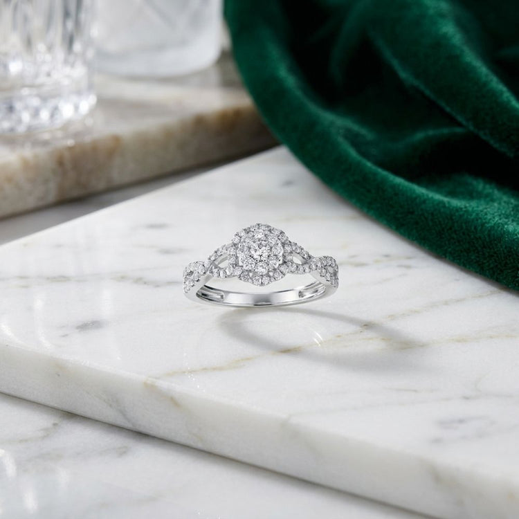 A 1/2 CTW Diamond Flower Bridal Ring in 14K gold rests on white marble beside green velvet fabric and a glass, creating an elegant scene.