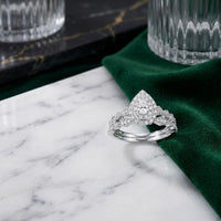 A 10k Gold Pear Diamond Halo Bridal Ring Set 3/8 Cttw, featuring a matching band, is displayed on a white marble surface beside green velvet fabric and ribbed glassware, creating an elegant bridal look.