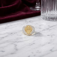 The 10k Gold 7/8 CTTW Diamond Cross Men’s Ring features a bold raised cross design, crafted in yellow gold and adorned with natural diamonds. It rests elegantly on a white marble surface, while a burgundy velvet cloth and crystal glass in the background impart an air of sophistication.