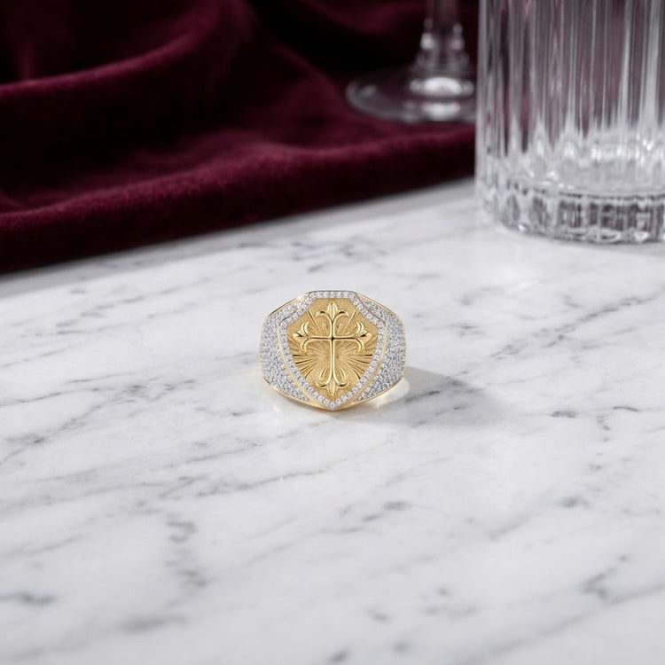 The 10k Gold 7/8 CTTW Diamond Cross Men’s Ring features a bold raised cross design, crafted in yellow gold and adorned with natural diamonds. It rests elegantly on a white marble surface, while a burgundy velvet cloth and crystal glass in the background impart an air of sophistication.