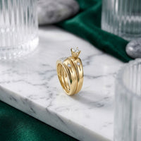 A 10k Gold Diamond Matching Wedding Ring Set sits gracefully on a white marble surface, encircled by clear glasses, smooth gray stones, and deep green fabric—an elegant scene that highlights the set’s timeless beauty.