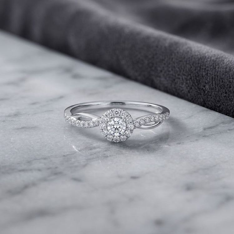 A dazzling 10k yellow gold round diamond halo promise ring, showcasing a brilliant round center stone surrounded by smaller diamonds totaling 1/5 cttw, rests gracefully on a white marble surface with a soft gray fabric in the background.