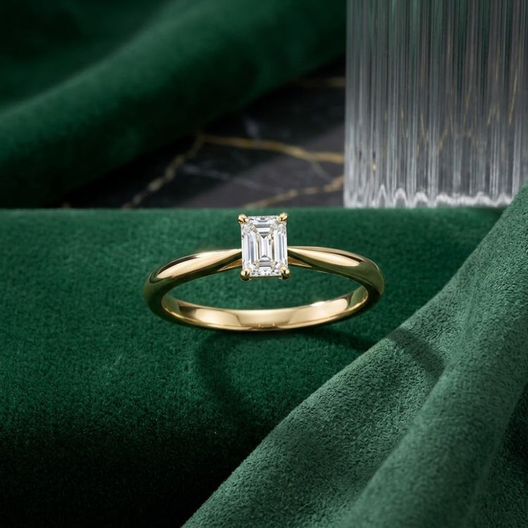 A stunning 1/4CT Diamond Emerald Solitaire Ring in 14K gold is displayed on green velvet fabric, with a ribbed glass object in the background, highlighting this elegant natural diamond solitaire.