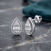 1/10 CTW Natural Diamond Pear Earrings in 10k Gold, featuring baguette and round diamonds in a teardrop shape, displayed on gray marble beside teal fabric.
