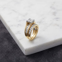 The 14K Gold Princess Diamond Bridal Ring Set 2 CTTW features two elegantly modern rings with dazzling embedded diamonds, including a breathtaking 2 carat center diamond. Both rings rest atop a pristine white marble surface, while a contrasting dark textured background enhances the brilliance and sophistication of this exquisite bridal set.