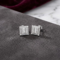A pair of 10K Gold Diamond Square Earrings 1/2 CTTW with a double halo design, showcased on a gray suede surface accented by marble and burgundy fabric in the background.