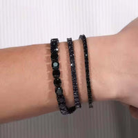A wrist displays three Black Moissanite Tennis Bracelets of different thicknesses, all set in 925 sterling silver, against a light striped background.