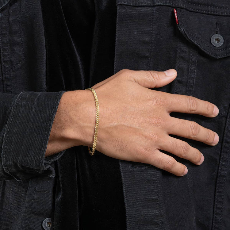 A person wearing a black denim jacket rests their hand on their chest, showcasing the Cuban Link Bracelet - 6mm in sleek yellow gold delicately adorning their wrist.