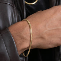 A close-up of a person wearing the Cuban Link Bracelet - 3mm in solid gold on their wrist, with a brown leather jacket sleeve and another gold chain necklace visible in the background.