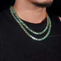 A person wears a sleeveless black top with two layered gold chains and the Green Moissanite Tennis Chain - 4mm, featuring large, sparkling blue-green gemstones around their neck.