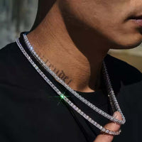 A close-up of a person wearing two sparkling 925 sterling silver chains—one is the Asscher Cut Moissanite Tennis Chain - 4mm—over a black shirt, with a neck tattoo visible and sunlight forming a star-shaped lens flare on the chain.