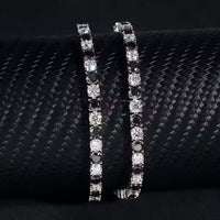 The Black Moissanite Tennis Bracelet - 4mm, crafted from 925 sterling silver with alternating black and white stones, is elegantly displayed on a textured black cylindrical surface for a striking contrast.
