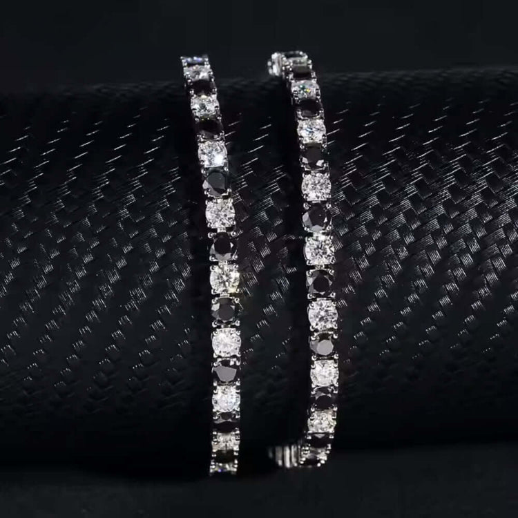 The Black Moissanite Tennis Bracelet - 4mm, crafted from 925 sterling silver with alternating black and white stones, is elegantly displayed on a textured black cylindrical surface for a striking contrast.