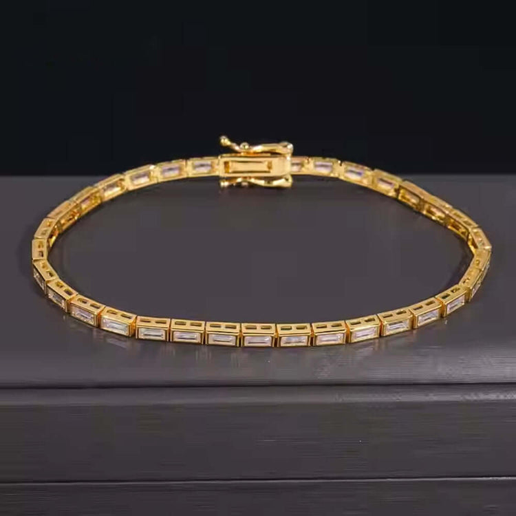 The Emerald Moissanite Tennis Bracelet - 4mm, featuring rectangular gold links and clear emerald-cut moissanite gemstones, is displayed on a dark, smooth surface against a black background.