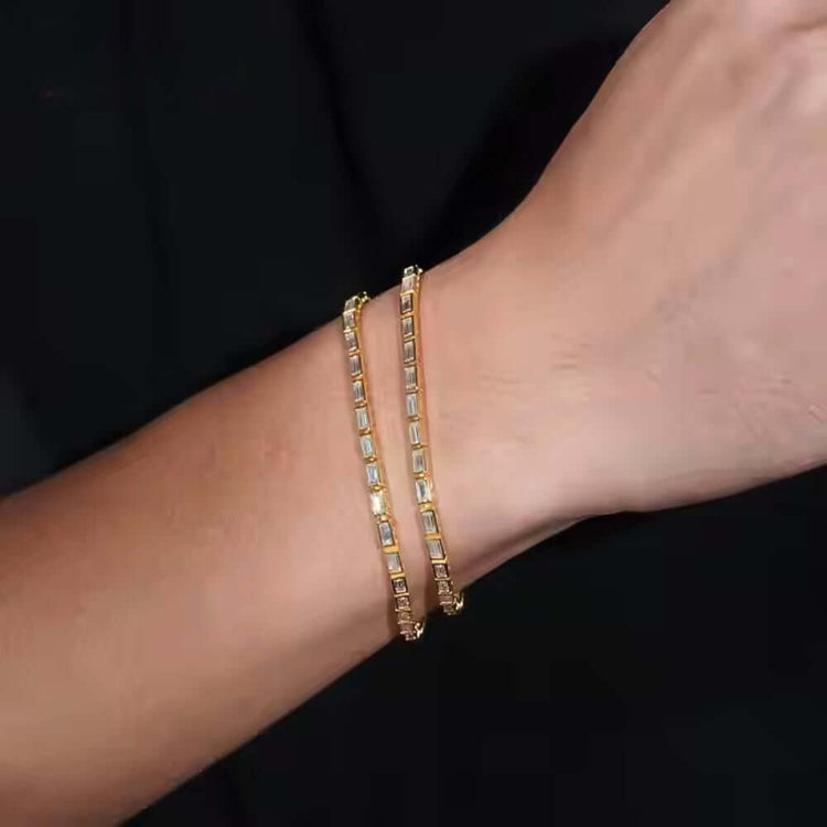 A close-up of a wrist wearing two Emerald Moissanite Tennis Bracelets - 4mm, featuring thin gold chains and emerald-cut moissanite stones, set against a black background.