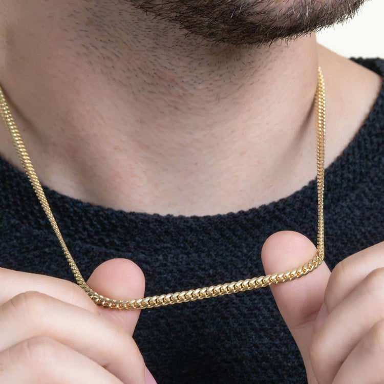 A person in a black sweater holds the Cuban Link Chain Solid Gold 4mm - Box Lock near their neck with both hands, highlighting the necklace and lower face against a plain background.