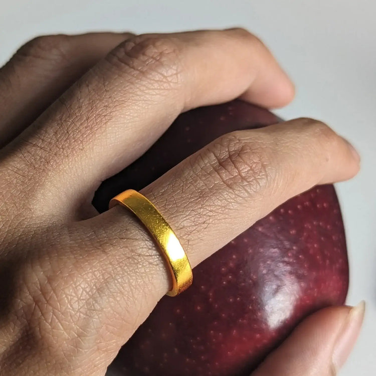 A hand wearing the 4mm Flat Band Ring - 24k holds a dark red apple against a light background.