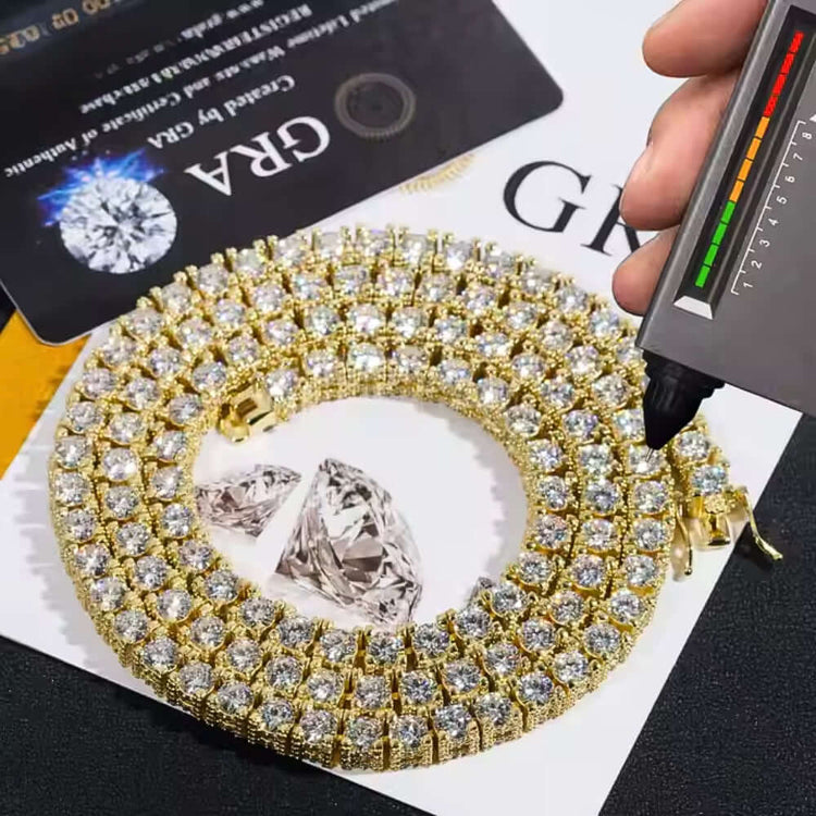 A Fully Ice Moissanite Tennis Chain - 5mm lies coiled on a surface next to two loose diamonds. A gemstone tester touches the chain, with certificates or cards visible in the background.