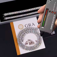 A hand tests an Iced Out Moissanite Tennis Chain (5mm) and loose stones with a diamond tester; a diamond bracelet and GRA certification card are also seen on the table.
