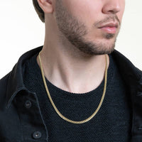 A man with a trimmed beard wears the Cuban Link Chain Solid Gold 5mm - Box Lock over a textured black shirt and black jacket, facing slightly to the side against a light background.