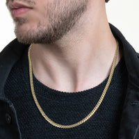 A man with a short beard wears the Cuban Link Chain Solid Gold 5mm - Box Lock over a black textured shirt and dark jacket. The focus is on the solid gold chain around his neck and his lower face; background is plain.