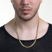 A man wearing a black shirt and the Cuban Link Chain Solid Gold 6mm - Box Lock necklace, shown from the shoulders up against a neutral background. His face is partially visible, highlighting trimmed facial hair.