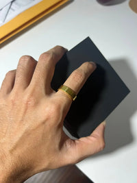 A hand wearing the 6mm Flat Band Ring -24k holds a small, plain black box on a white surface, with an envelope and paper partially visible in the background.
