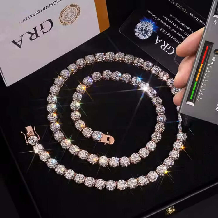A hand tests the Rose Gold Moissanite Tennis Chain- 8mm with a diamond tester; the sparkling necklace and matching bracelet, set with round-cut stones, are displayed in a black box beside a GRA certificate.