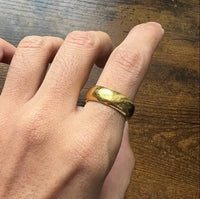 A hand wears the 8mm Band Ring -24k, a thick and shiny comfort-fit 24k gold ring, on the middle finger while resting on a wooden surface.