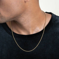 Close-up of a person wearing Beads Chains, a delicate necklace in 14K gold, over a black velvet shirt. Only their lower face, neck, and upper chest are shown.