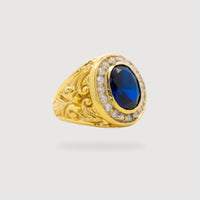 The Blue Stone Signet Ring in Solid Gold features a large oval blue gemstone at the center, bordered by small clear crystals and intricate detailing, displayed on a plain light background.