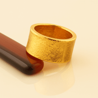 The 13mm 1oz Flat Band Ring - 24k, with a wide textured minimalist gold design, rests on a glossy dark red and black cylinder against a light beige background.