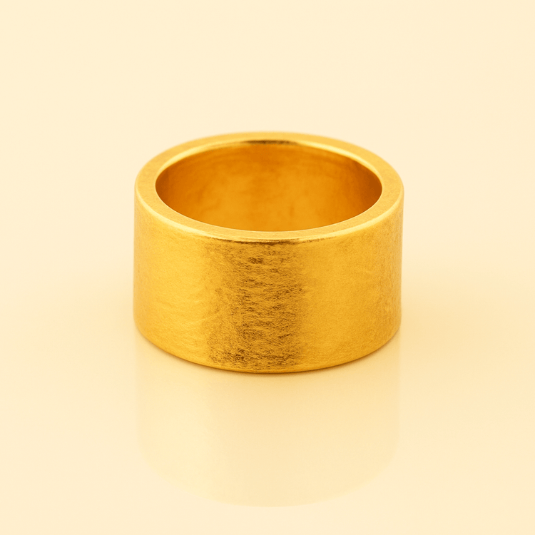 The 13mm 1oz Flat Band Ring - 24k, a wide matte gold band, stands upright on a glossy light beige surface with a matching minimal background for a warm, sophisticated look.