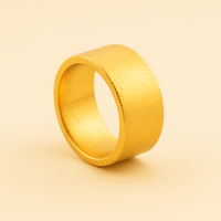 The 13mm 1oz Flat Band Ring - 24k stands upright on a matching light beige background, highlighting its wide, textured band and minimalist gold design.