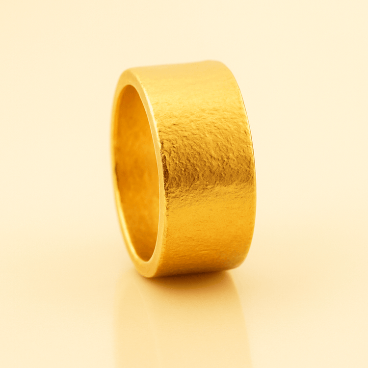 The 13mm 1oz Flat Band Ring - 24k stands upright on a smooth, light beige surface, showcasing its wide, textured minimalist gold design with a matte finish for a simple and elegant display.
