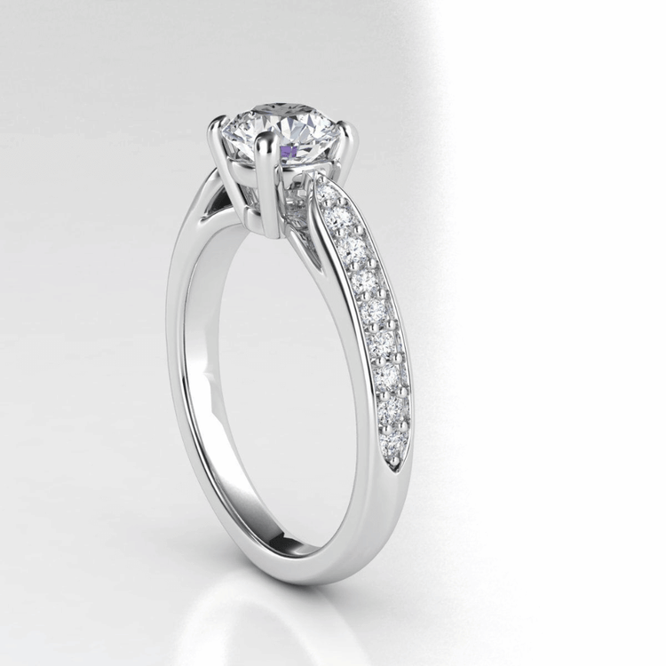 The Classic Round-Cut Lab Diamond Engagement Ring with Side Pave Details features a brilliant round lab diamond in four prongs, accented by sparkling side pave stones, elegantly shown on a white background.