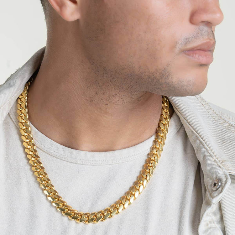 A man in a light jacket and t-shirt wears the Cuban Link Chain 12mm - 14k Gold Bonded around his neck. His face is partly visible against a plain, light background.