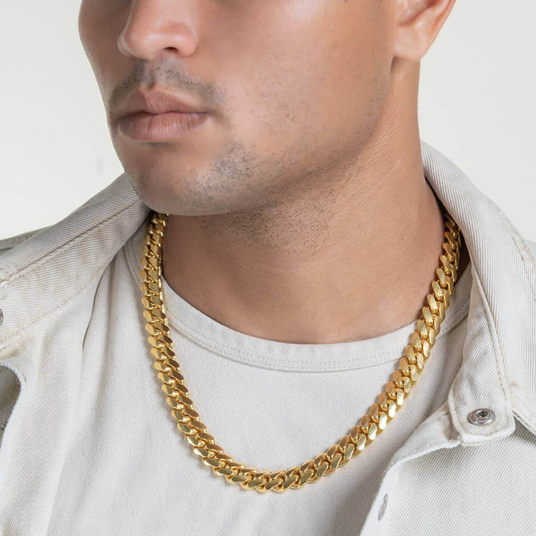 A person wearing a cream jacket and t-shirt is shown from the neck down, highlighting the Cuban Link Chain 12mm - 14k Gold Bonded necklace, which rests around their neck against a light background.