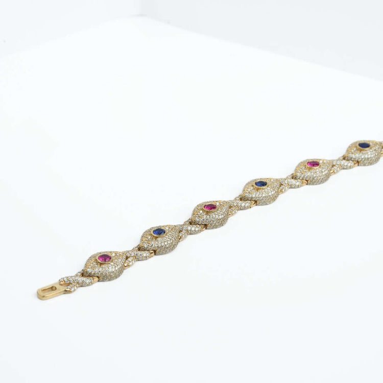 The Diamond Evil Eye Gemstone Bracelet in 10k gold features a wavy design, encrusted with small white stones and alternating blue and pink gemstones, displayed on a white surface.